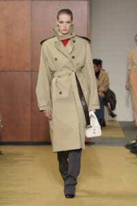 Model walking a runway in a beige double-breasted trench coat with a belt, red top peeking through, and gray trousers with a white handbag by her side.