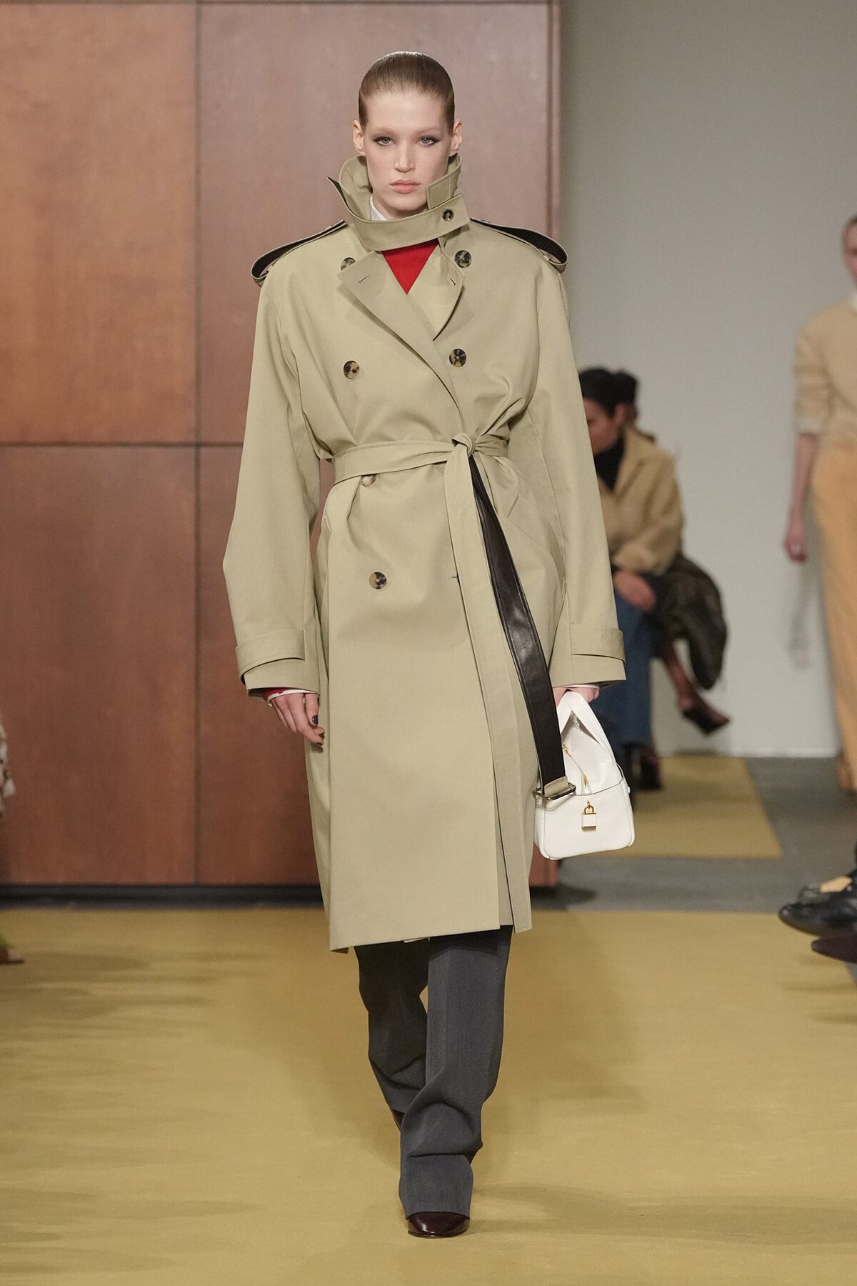 Model walking a runway in a beige double-breasted trench coat with a belt, red top peeking through, and gray trousers with a white handbag by her side.