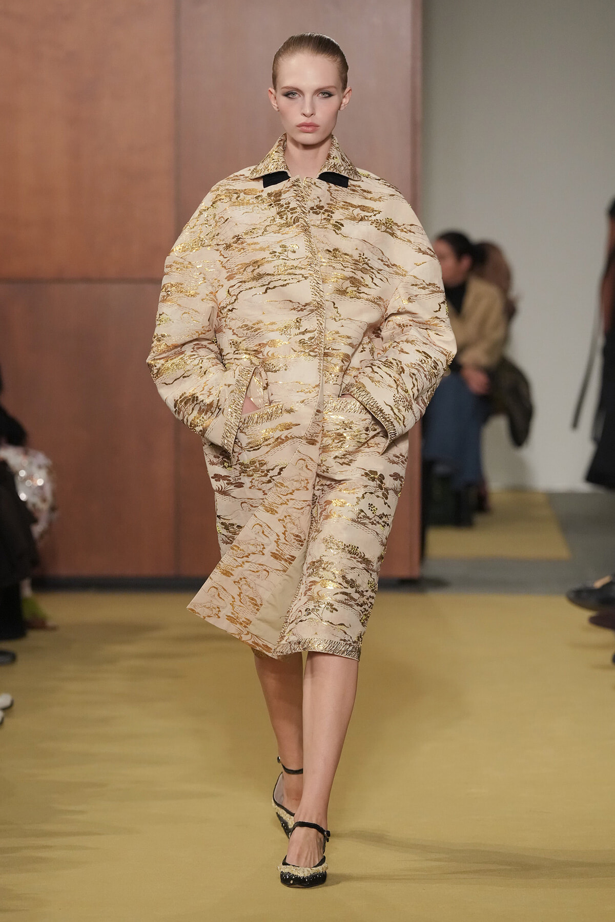 Model walks the runway in a beige brocade suit with gold metallic patterns, hands in pockets, neutral expression.