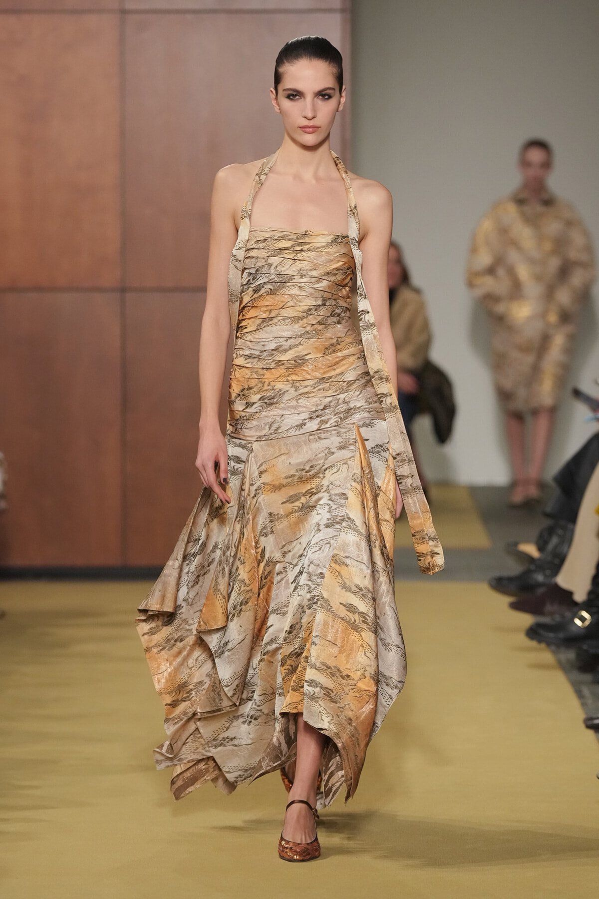Model on a runway wearing a strapless, tan and gold printed halter dress with layered, flowing skirt and slit, brown sandals.