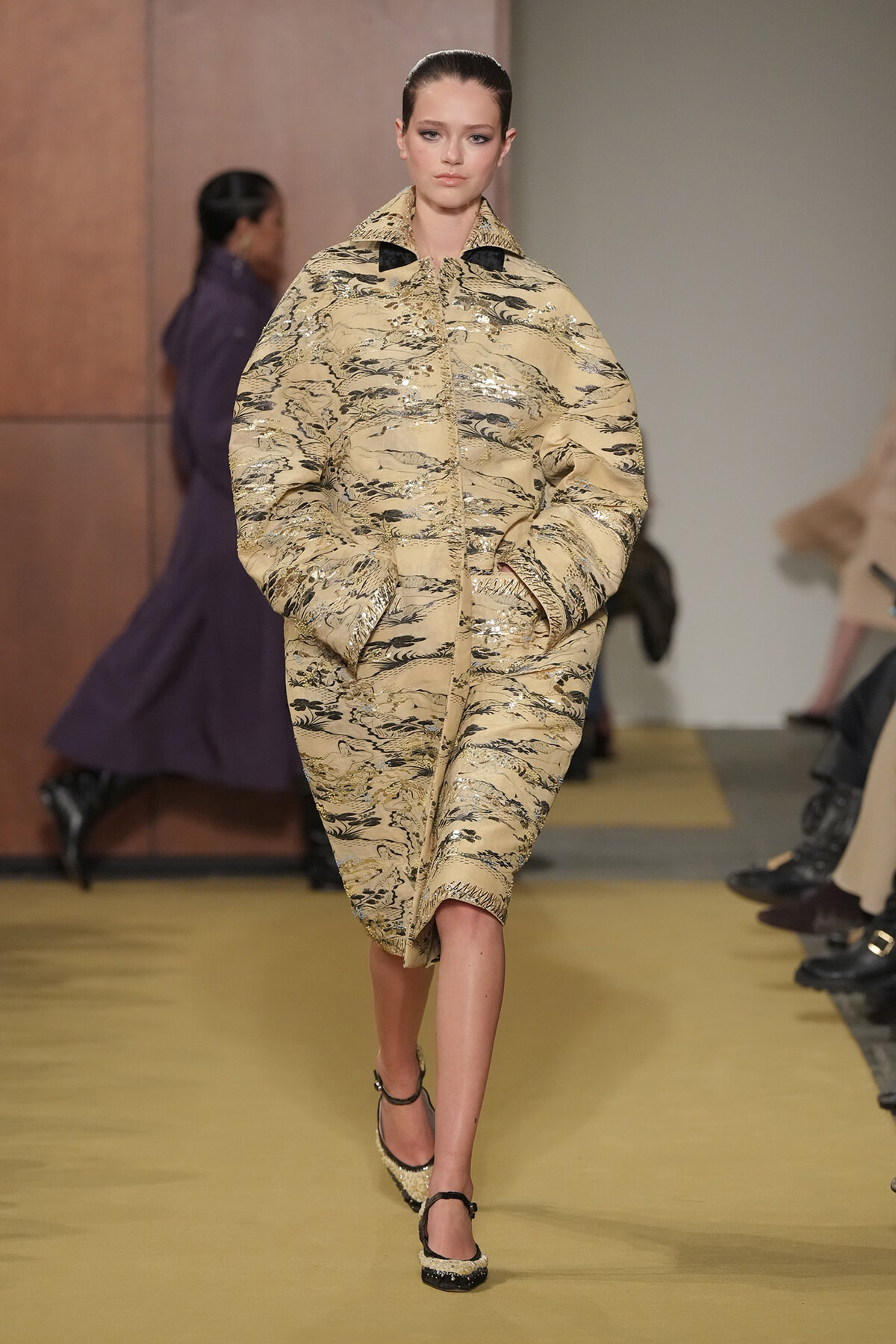 Model walking the runway in an oversized beige coat with metallic abstract patterns, hands in pockets.