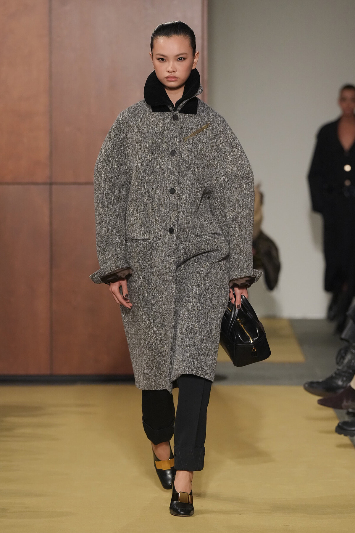 Model on runway wearing a voluminous grey coat with black collar, black pants, and black handbag.