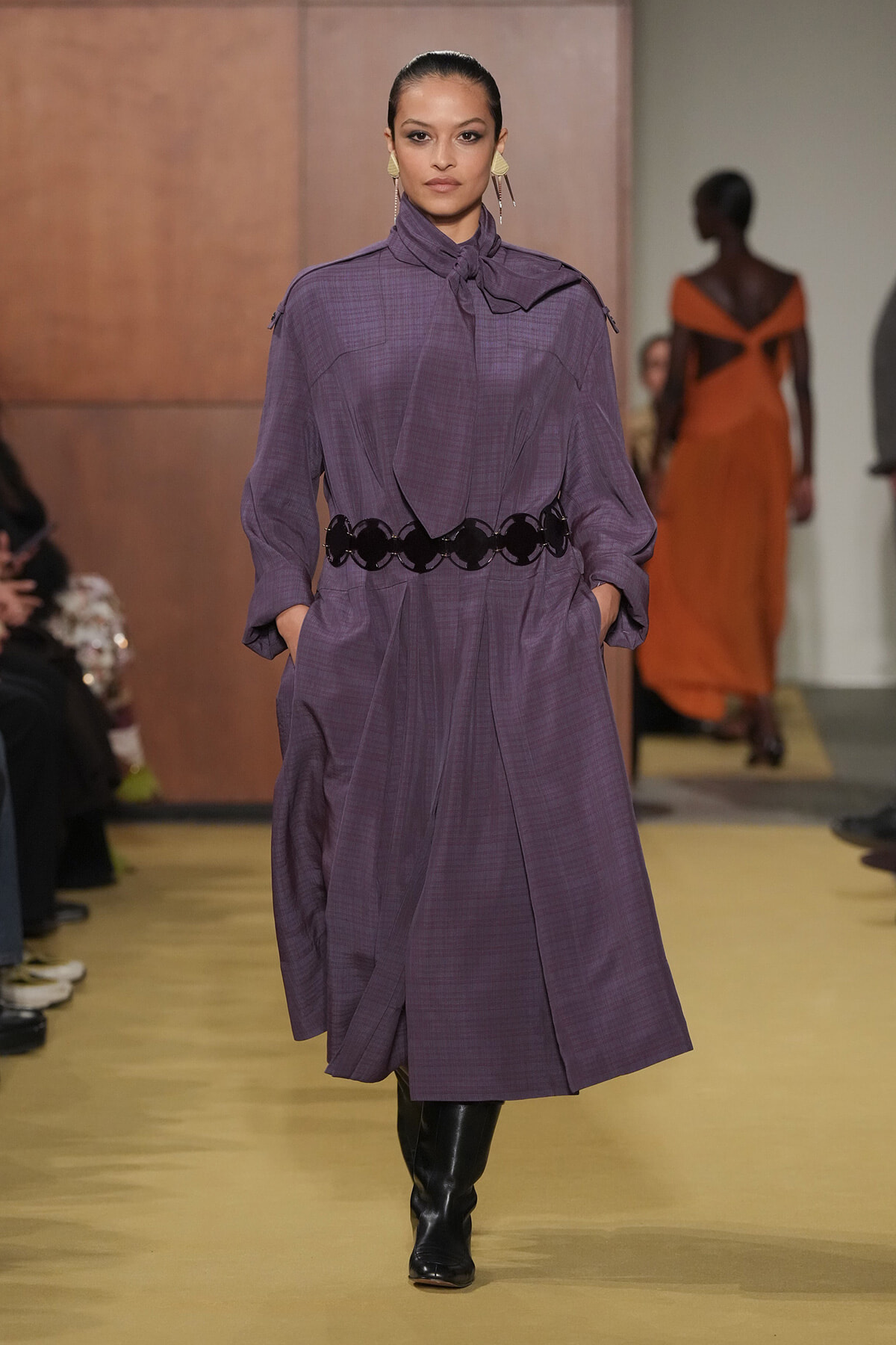 Model walking runway in a purple long-sleeve dress with a gathered neck scarf, cinched at the waist with a black circular belt, hands in pockets, black boots.
