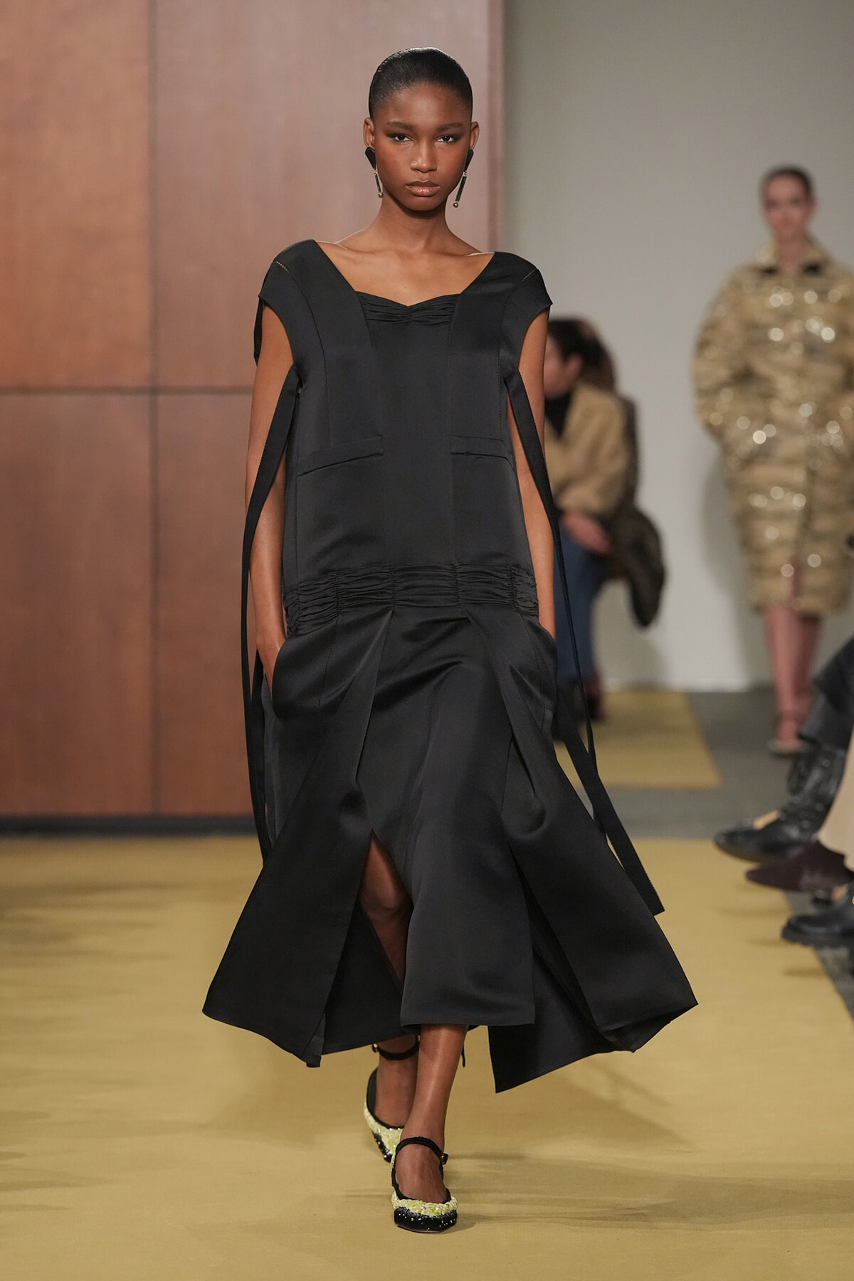 Model walking the runway in a black, structured midi dress with long cape-like sleeves and a ruched waist.