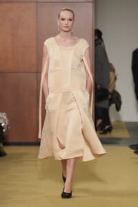 Model walking runway in a pale peach-toned dress with cape-like sleeves and a gathered waist, showing a front slit and black flats.