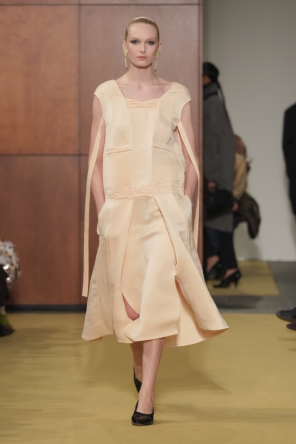 Model walking runway in a pale peach-toned dress with cape-like sleeves and a gathered waist, showing a front slit and black flats.