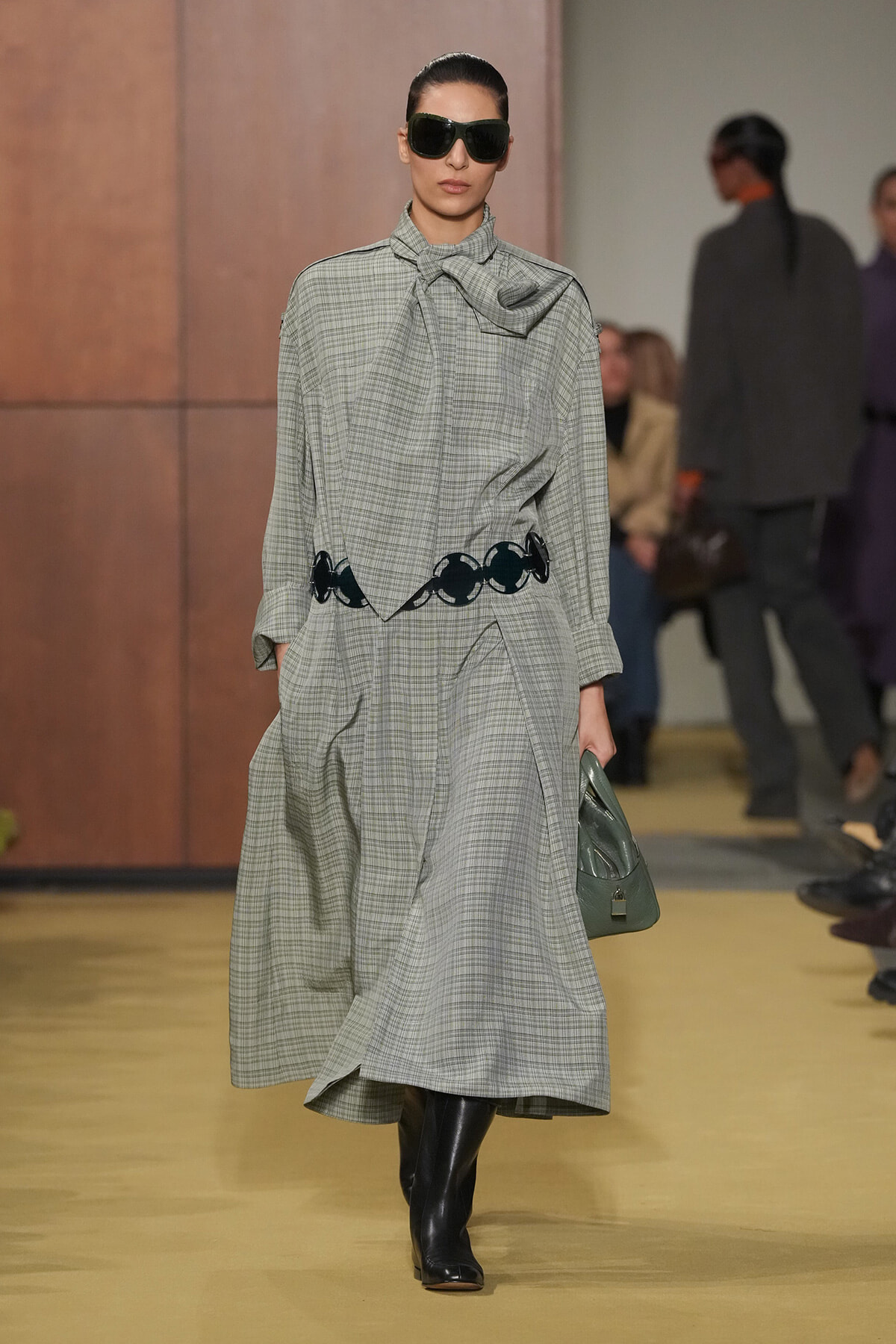 Model walks a runway in a gray checkered long dress with a large necktie bow, black belt circles, and black knee boots, carrying a green handbag and wearing oversized sunglasses