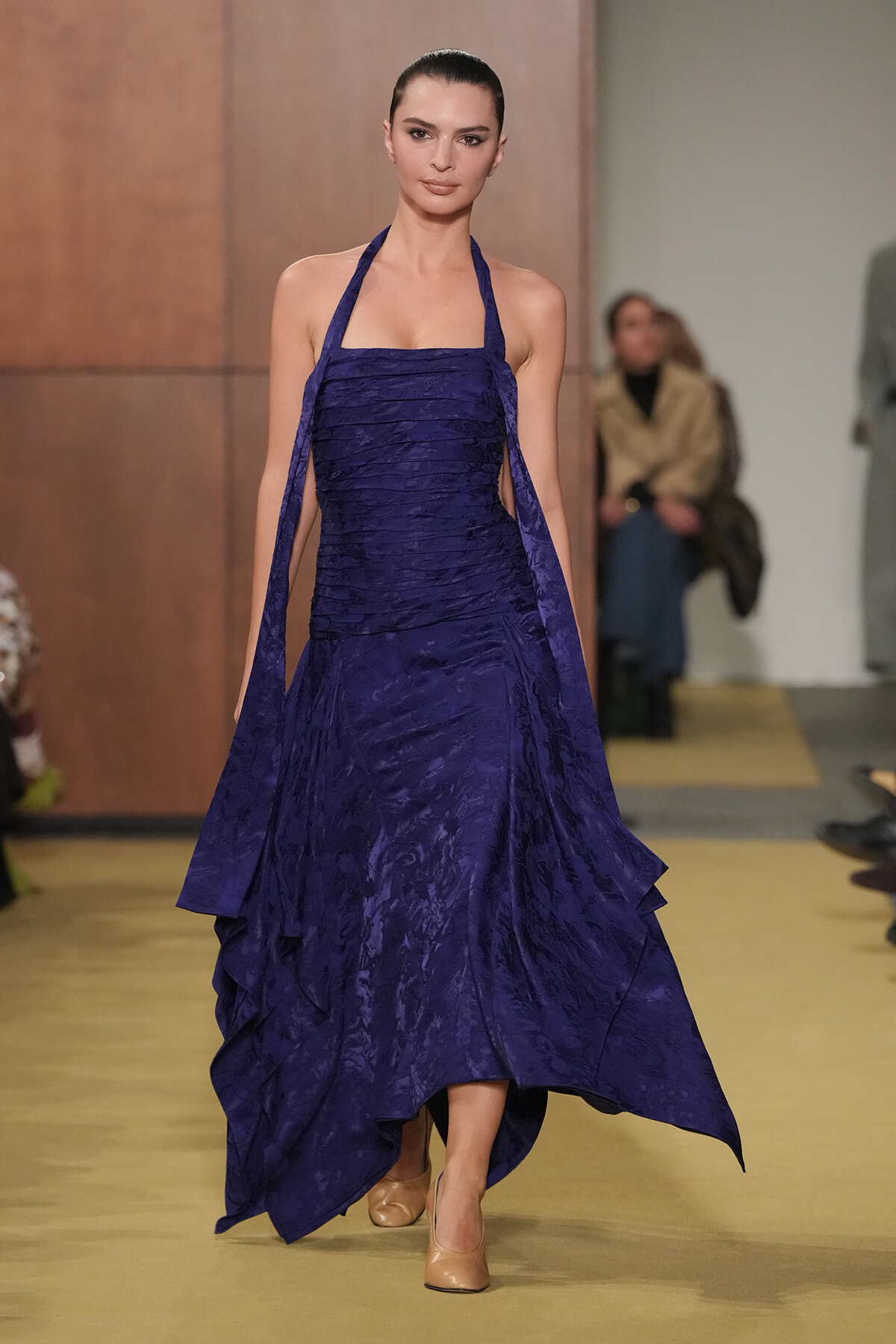 Model walks a purple halter-neck gown with a layered, flowing skirt on a runway.