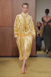 Model walks the runway in a gold-yellow patterned velvet suit with a draped scarf and matching belt, on a yellow-toned catwalk surface.