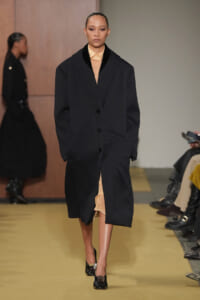 Runway model in an oversized black coat over a peach dress, walking on a yellow fashion show runway.