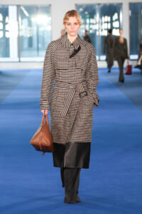 Model walking a blue runway in a beige-black checkered trench with a belt, carrying a brown handbag.