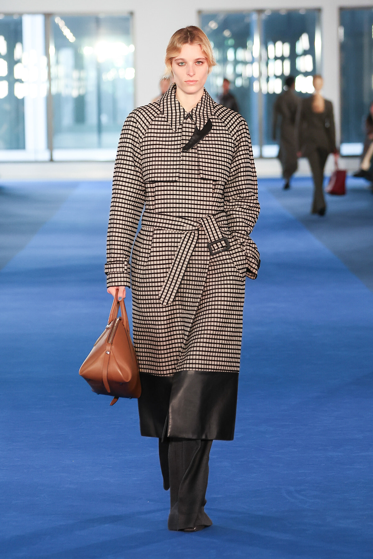 Model walking a blue runway in a beige-black checkered trench with a belt, carrying a brown handbag.