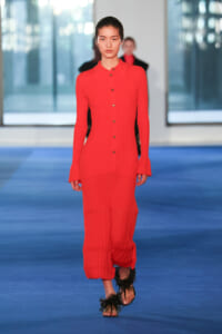 Model on a blue runway wearing a bright red ribbed knit jumpsuit with a button-down front and flared cuffs.