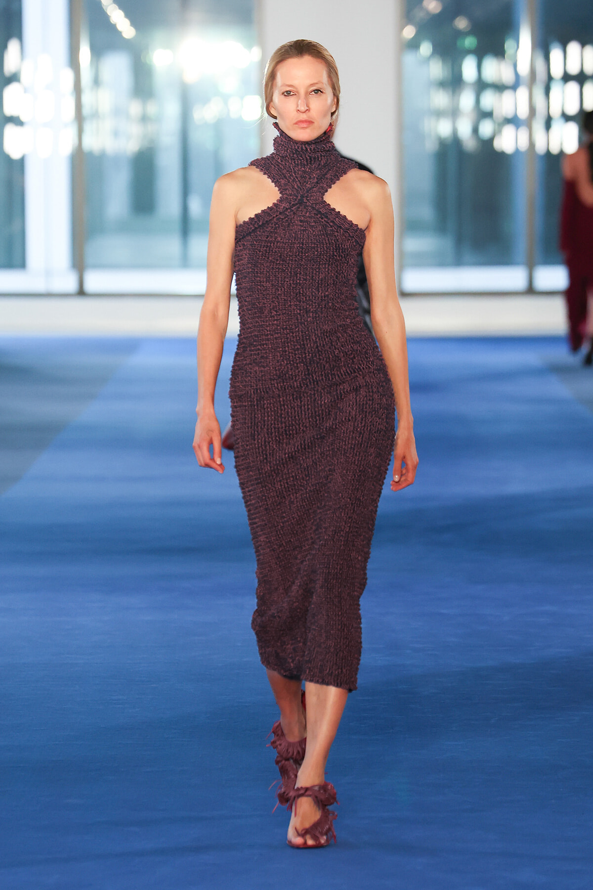 Model walking down a runway in a textured burgundy halter dress and matching heeled sandals.
