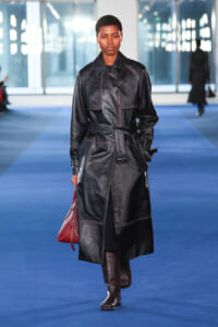 Model walks a blue runway in a long black leather trench coat with a belt, brown knee-high boots, and a red handbag.