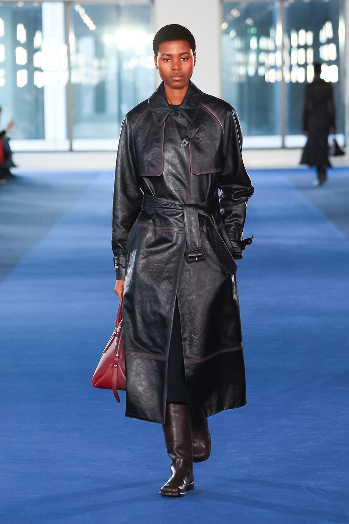 Model walks a blue runway in a long black leather trench coat with a belt, brown knee-high boots, and a red handbag.