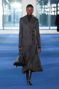 Model on a blue runway wearing a dark gray double-breasted blazer and matching long skirt, carrying a black handbag.