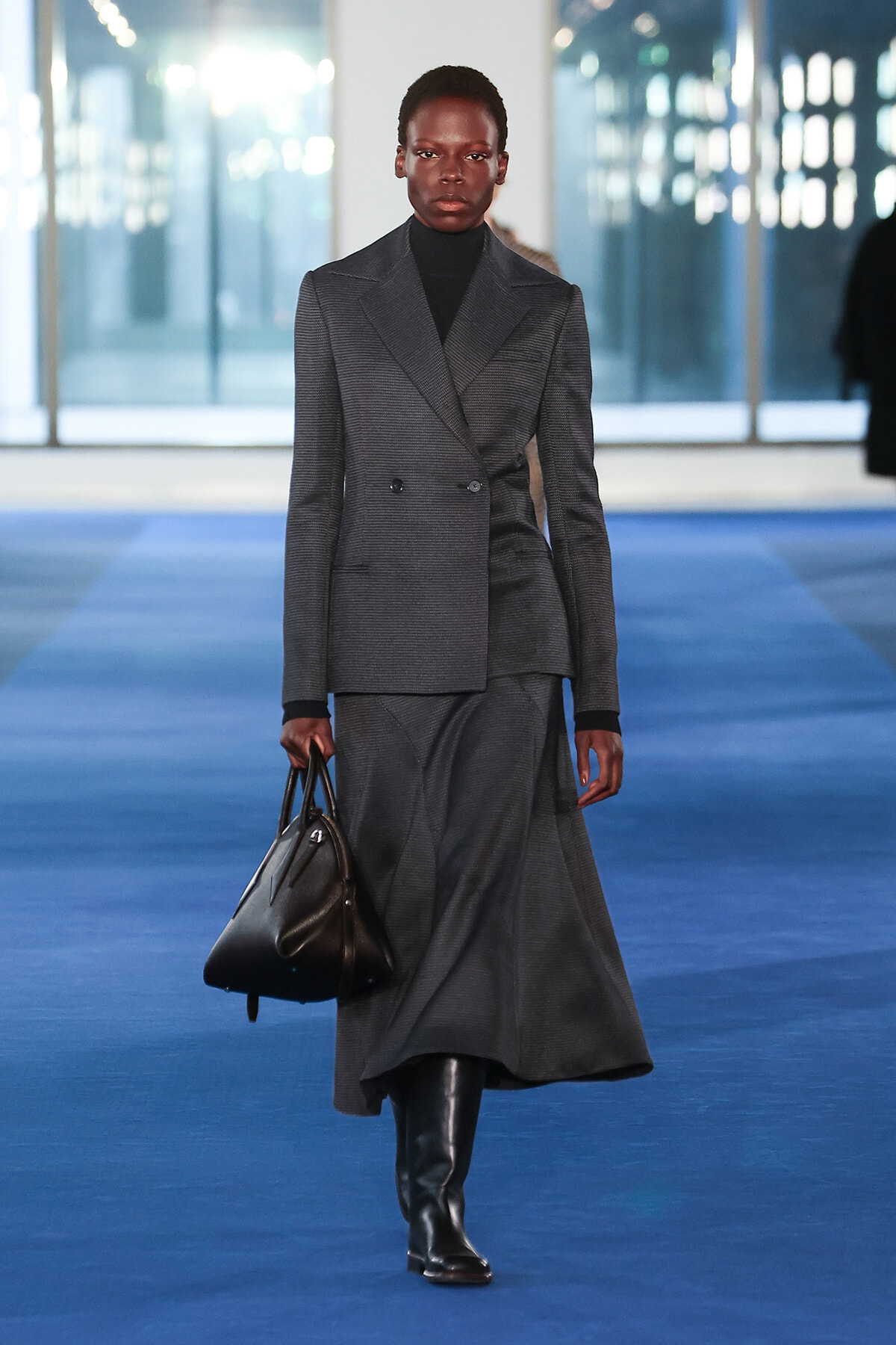 Model on a blue runway wearing a dark gray double-breasted blazer and matching long skirt, carrying a black handbag.