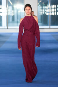 Female model walks the runway in a burgundy textured, off-shoulder halter-neck gown with long sleeves on a blue catwalk.