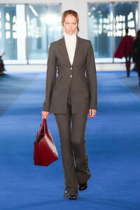 Female model walking a runway in a tailored gray suit with a white turtleneck, carrying a burgundy handbag on a blue catwalk.