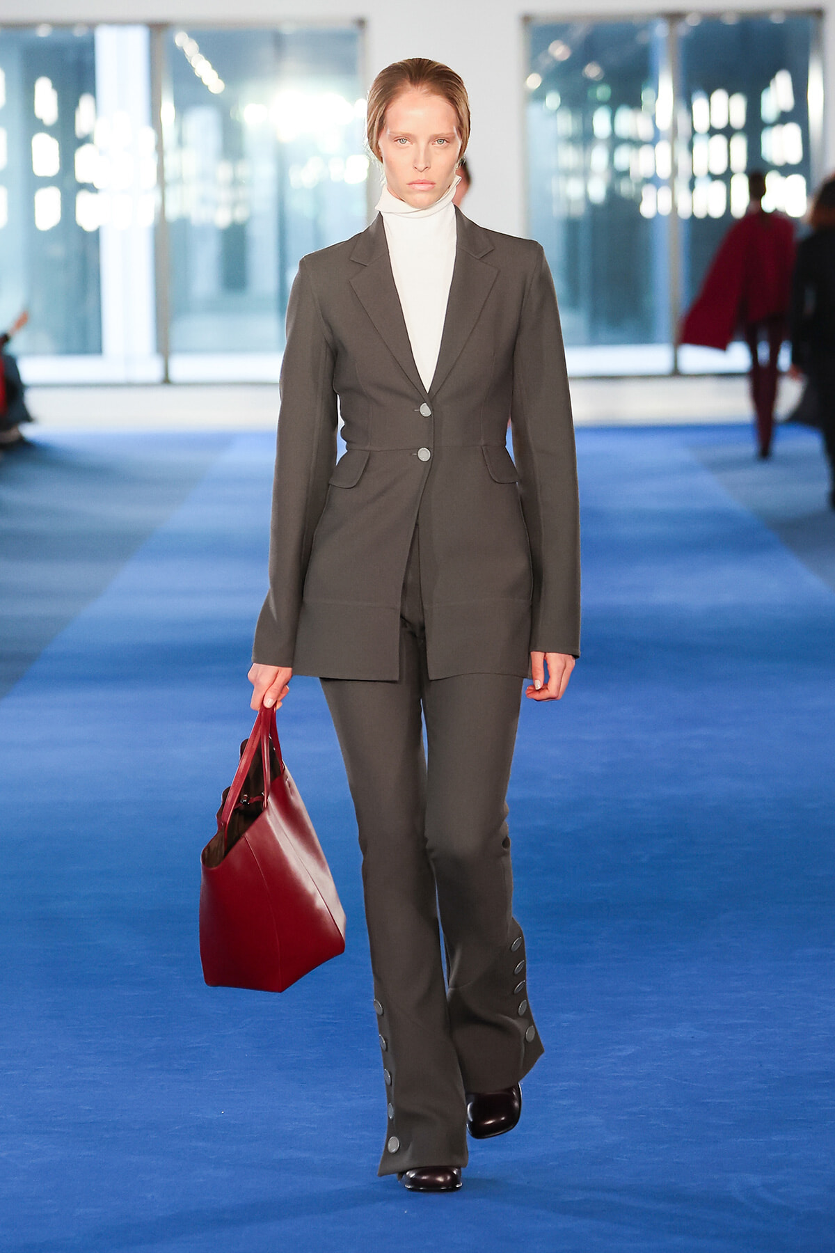 Female model walking a runway in a tailored gray suit with a white turtleneck, carrying a burgundy handbag on a blue catwalk.