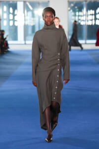 Model walks a blue runway in a gray turtleneck-style dress with a wrap waist and side buttons.