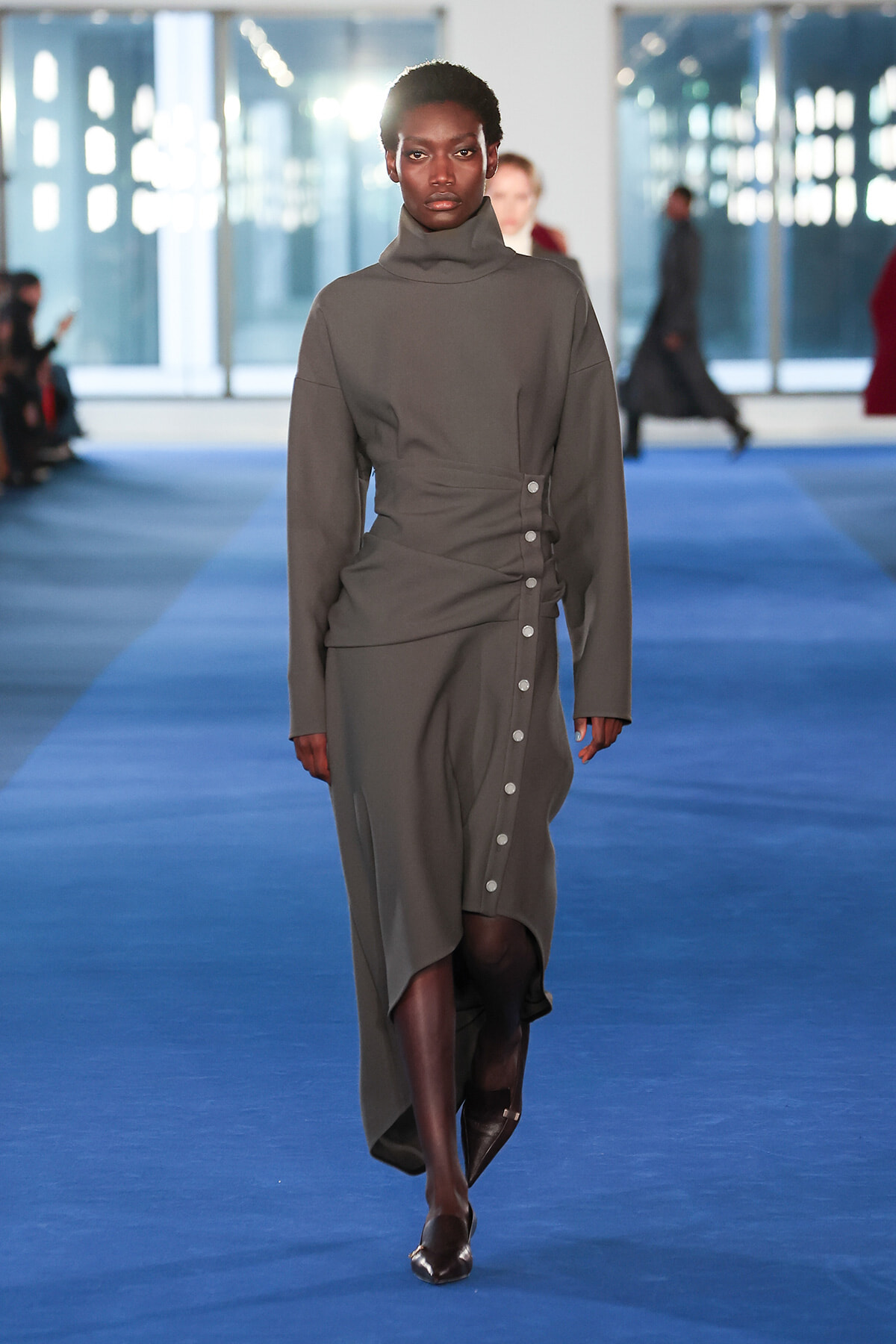 Model walks a blue runway in a gray turtleneck-style dress with a wrap waist and side buttons.