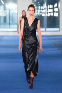 Model walking down a blue runway in a black satin wrap dress and brown ankle boots.