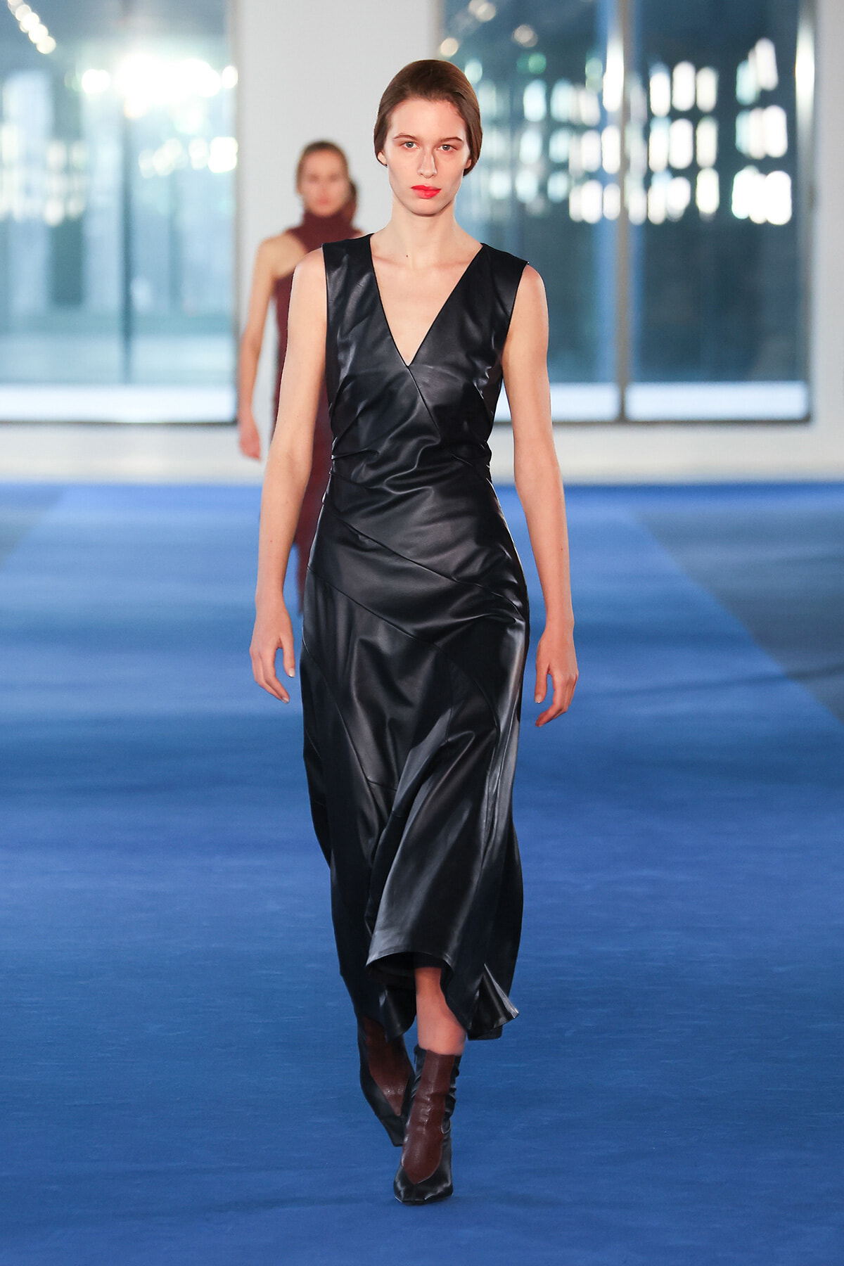 Model walking down a blue runway in a black satin wrap dress and brown ankle boots.