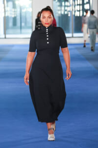 Model walks the blue runway in a knee-/ankle-length black polo dress with short sleeves and a row of white buttons at the neck, silver heels visible.