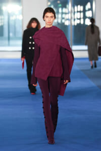 Model walks a runway in a burgundy cape-style top with matching pants and side-button detailing.