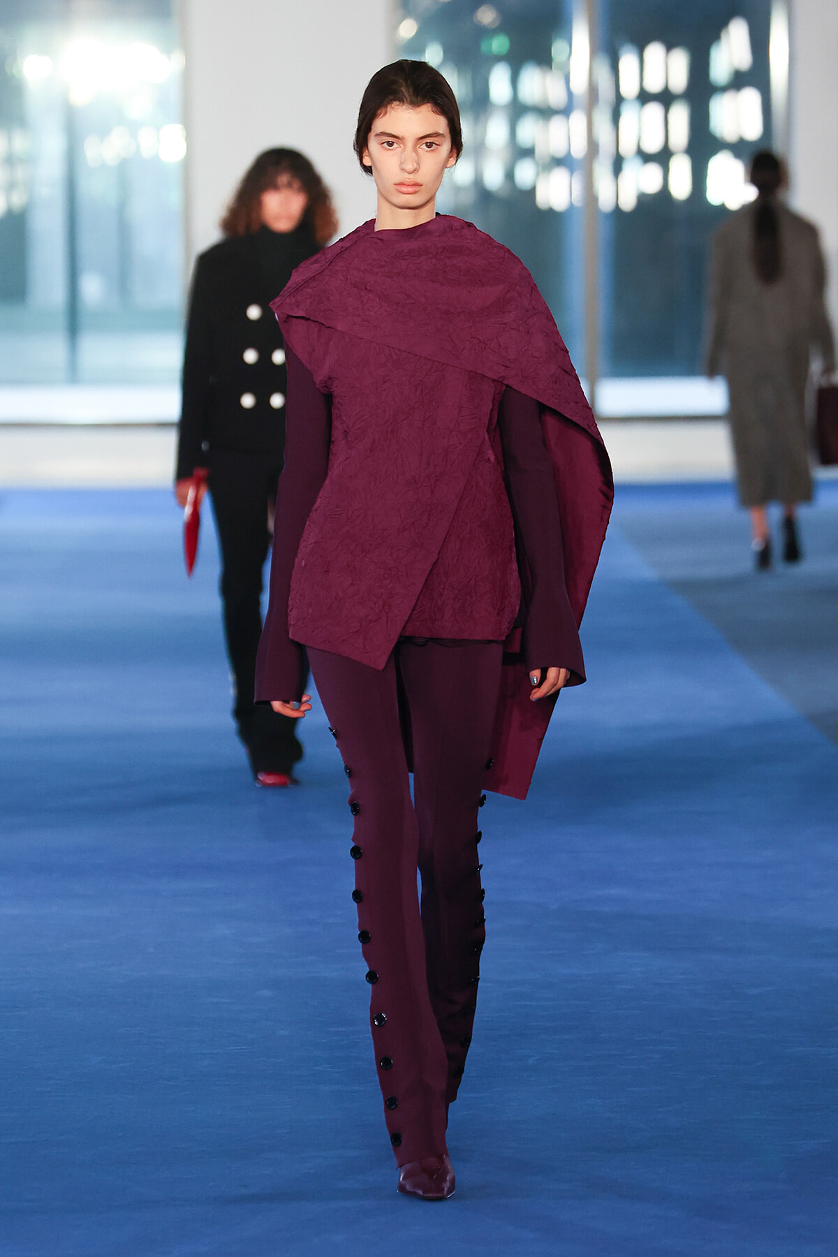 Model walks a runway in a burgundy cape-style top with matching pants and side-button detailing.