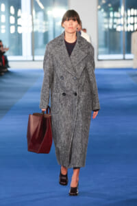 Model walking down a blue fashion runway in a long gray double-breasted coat, carrying a burgundy tote bag and wearing dark shoes.