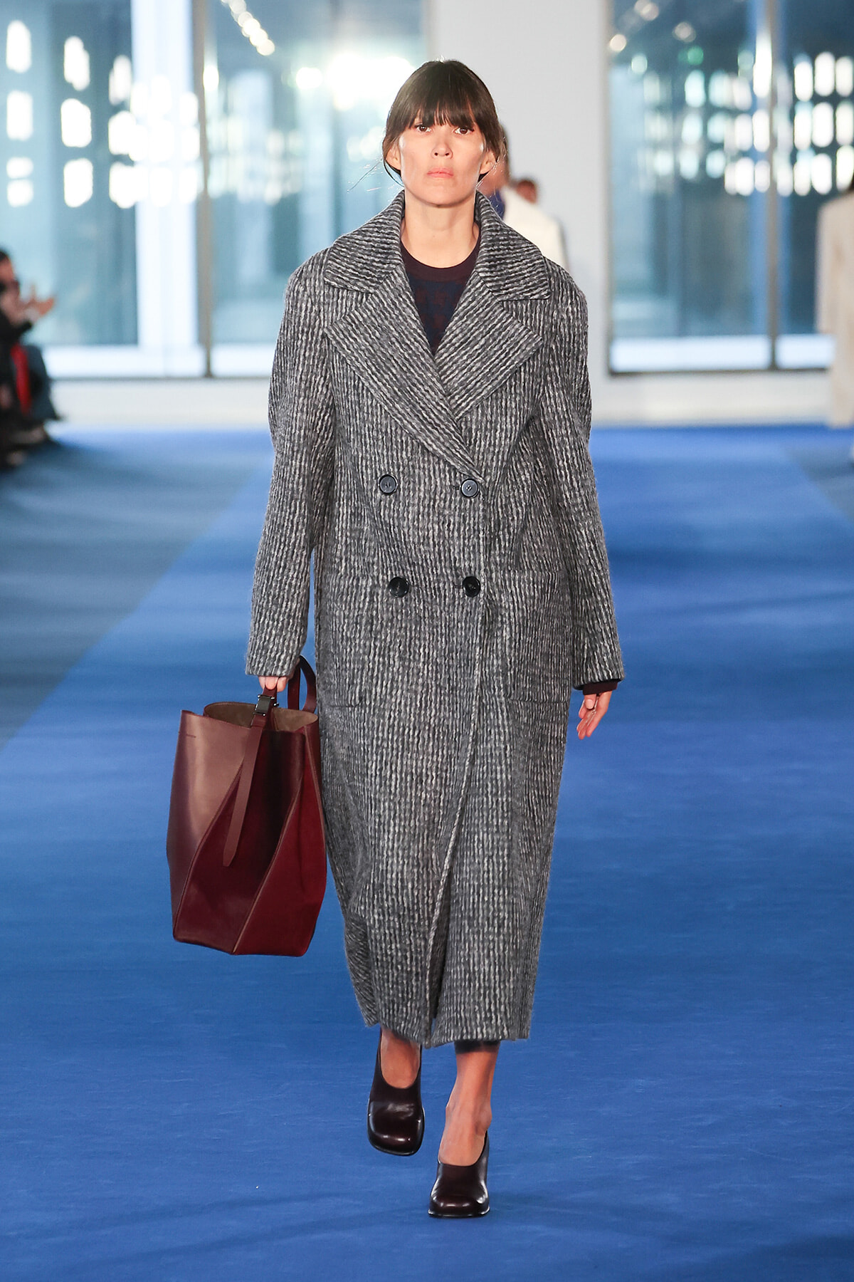 Model walking down a blue fashion runway in a long gray double-breasted coat, carrying a burgundy tote bag and wearing dark shoes.