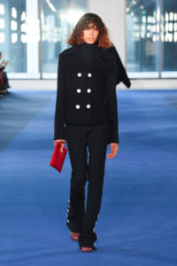 Model walks a blue runway wearing a navy double-breasted jacket with six white buttons, black trousers, and a red clutch.