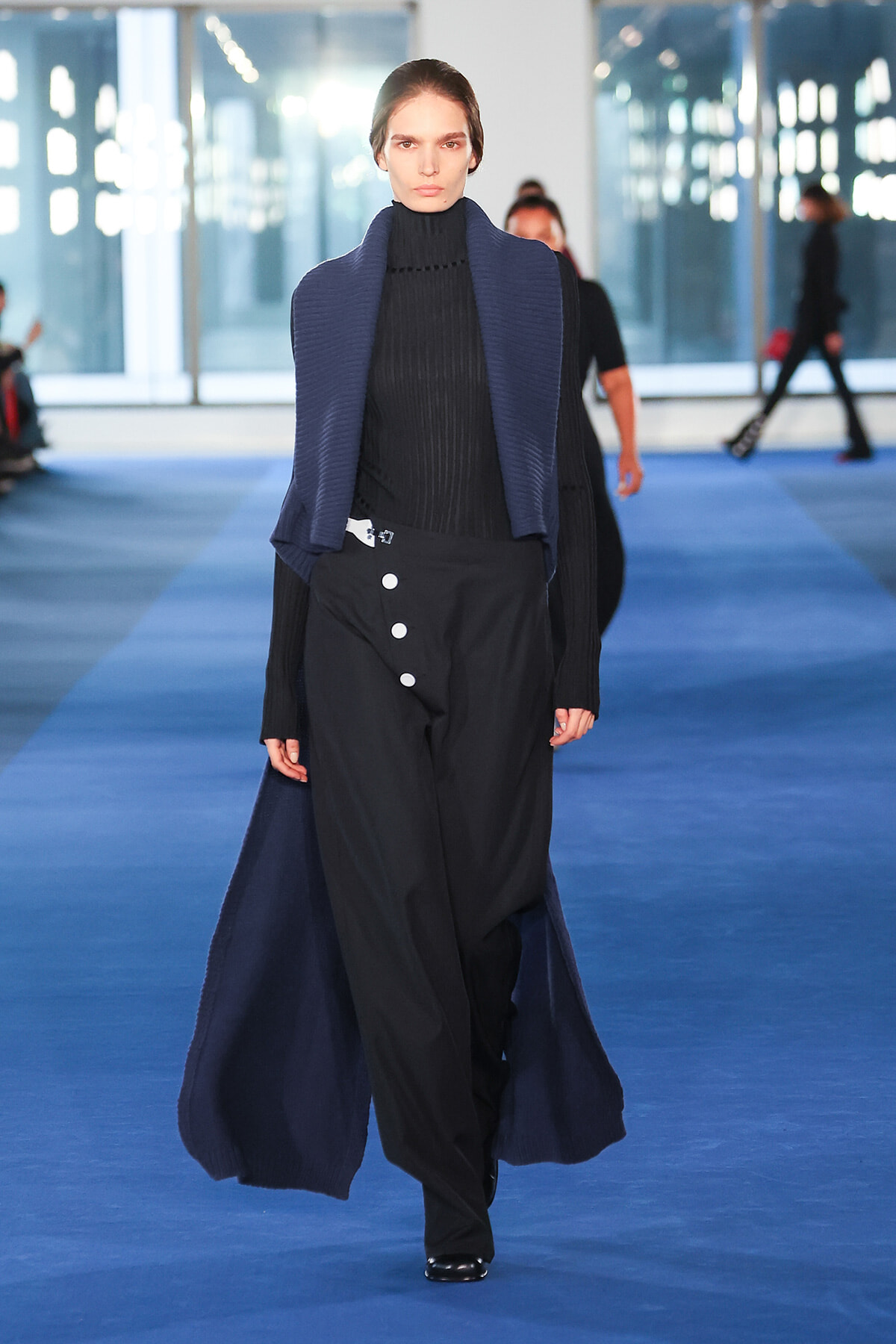 Model walking a blue runway in a black turtleneck, oversized navy cardigan, and high-waisted black pants with large white buttons.