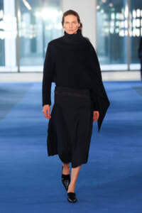Model walking a blue runway in an all-black outfit: oversized turtleneck and layered midi skirt with black shoes.