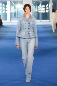 Female model wearing a light-wash denim suit (peplum jacket and matching jeans) walking a blue runway at a fashion show.