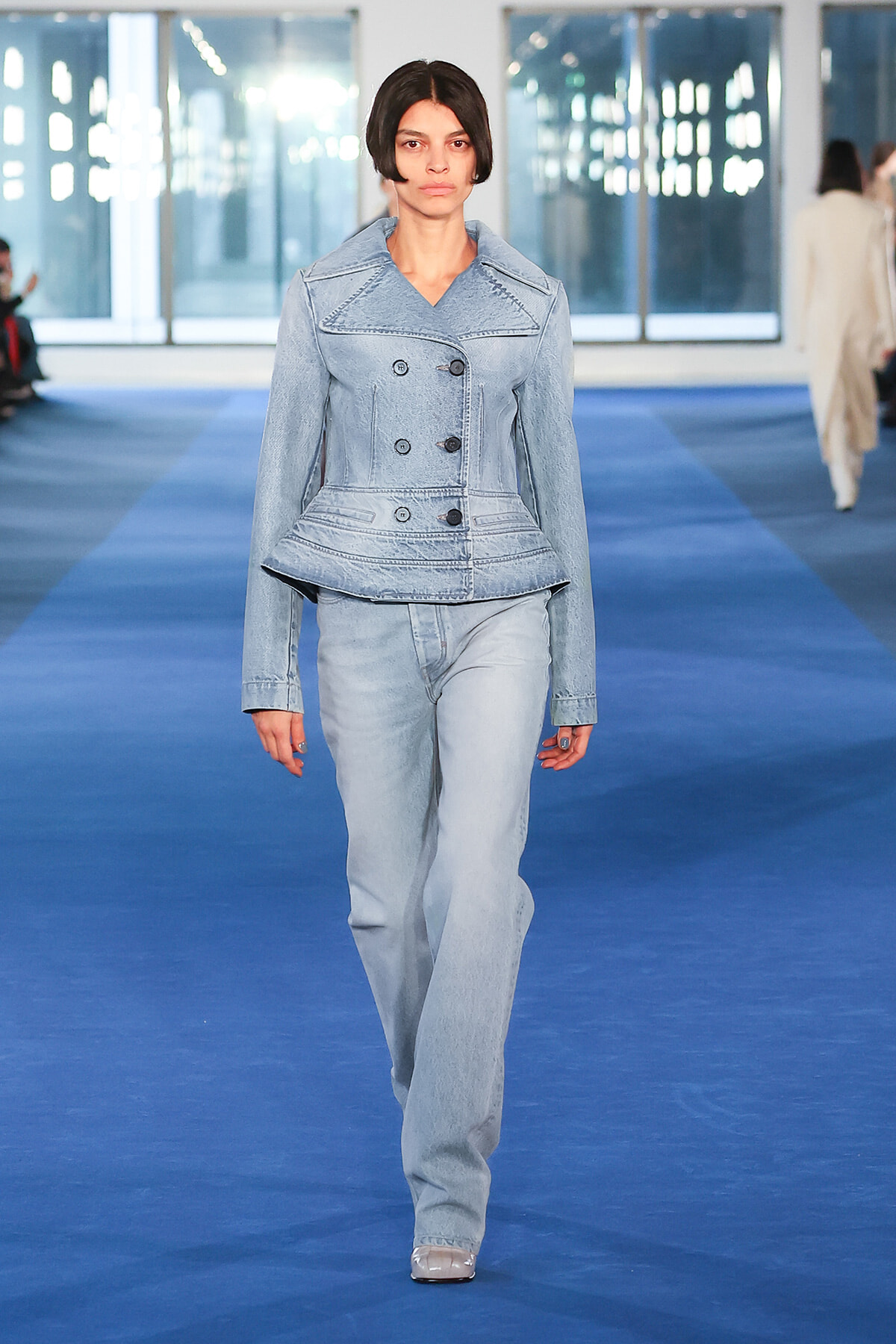Female model wearing a light-wash denim suit (peplum jacket and matching jeans) walking a blue runway at a fashion show.