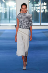 Model walking a fashion runway in a blue-toned scene, wearing a geometric navy-and-white printed top and a light gray pleated midi skirt with an asymmetric wrap detail.