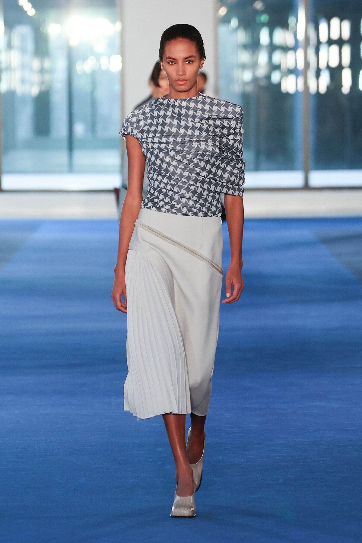 Model walking a fashion runway in a blue-toned scene, wearing a geometric navy-and-white printed top and a light gray pleated midi skirt with an asymmetric wrap detail.