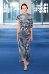 Female model walking a blue runway in a blue-gray geometric-patterned jumpsuit with a one-shoulder draped sleeve and neutral shoes.
