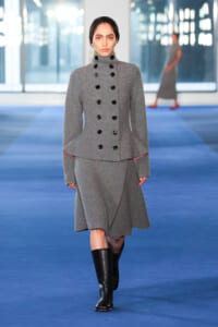 Model walking a runway in a gray double-breasted wool coat with black buttons and knee-high black boots on a blue catwalk floor.