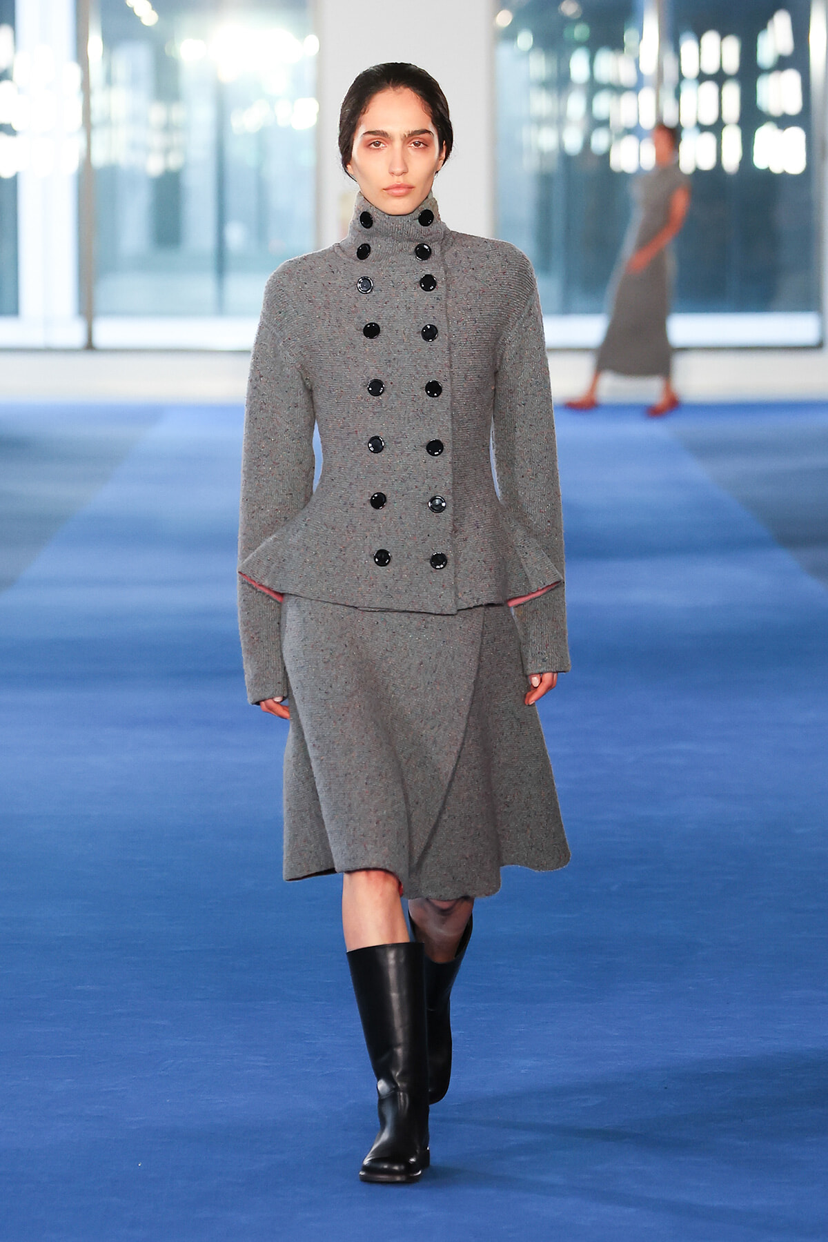 Model walking a runway in a gray double-breasted wool coat with black buttons and knee-high black boots on a blue catwalk floor.