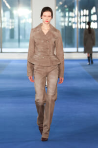 Model walking the runway in a taupe double-breasted suit with wide lapels and matching trousers.