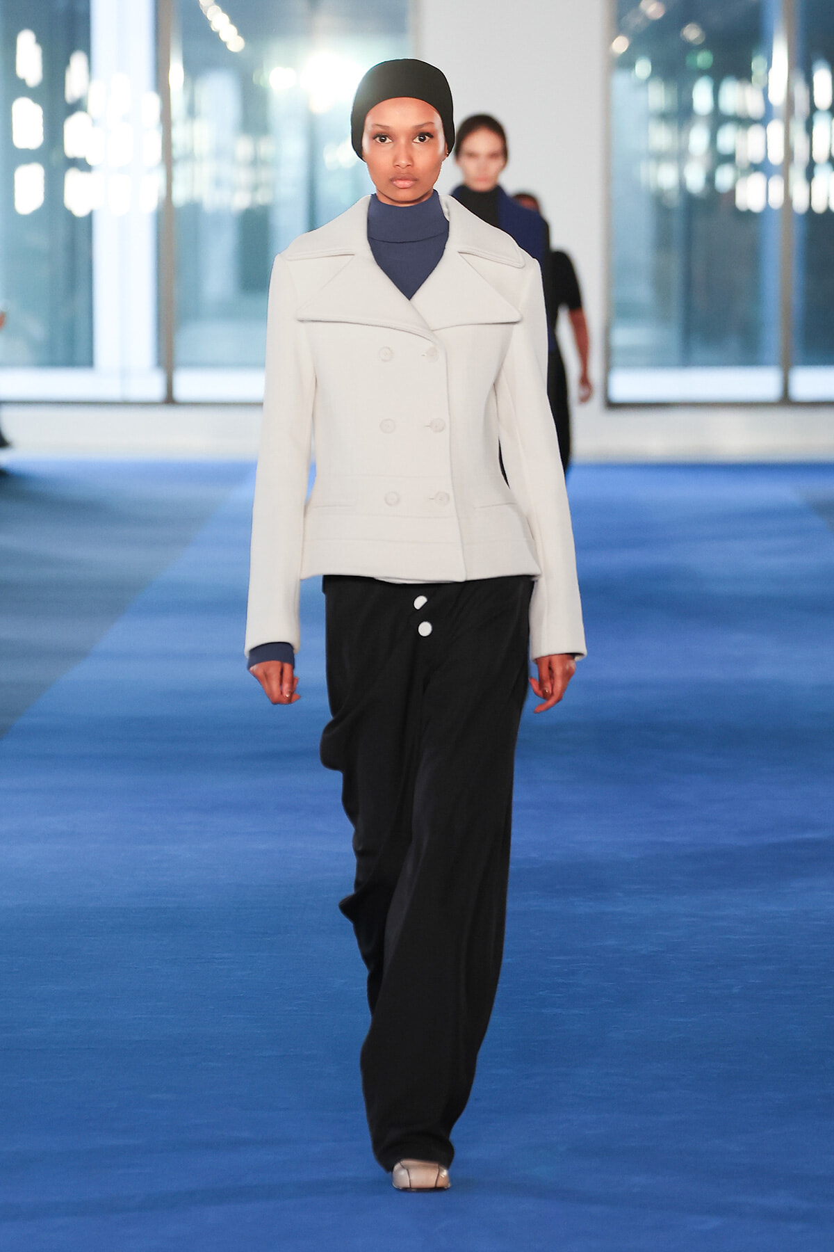 Model walking a blue runway in a white double-breasted jacket, black wide-leg pants, and a black headscarf.