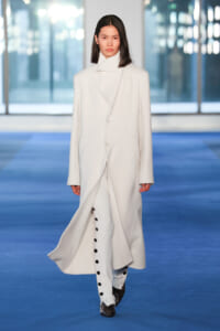 Model walking the runway in a long white wrap coat over matching white pants with a row of black buttons, on a blue catwalk backdrop.