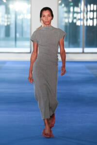 Runway model walking in a gray textured dress and pink fringed shoes on a blue catwalk.