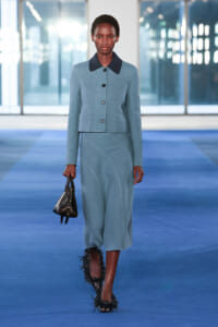 Model on a blue runway wearing a light blue cropped jacket with a dark collar and matching skirt, carrying a small black handbag.