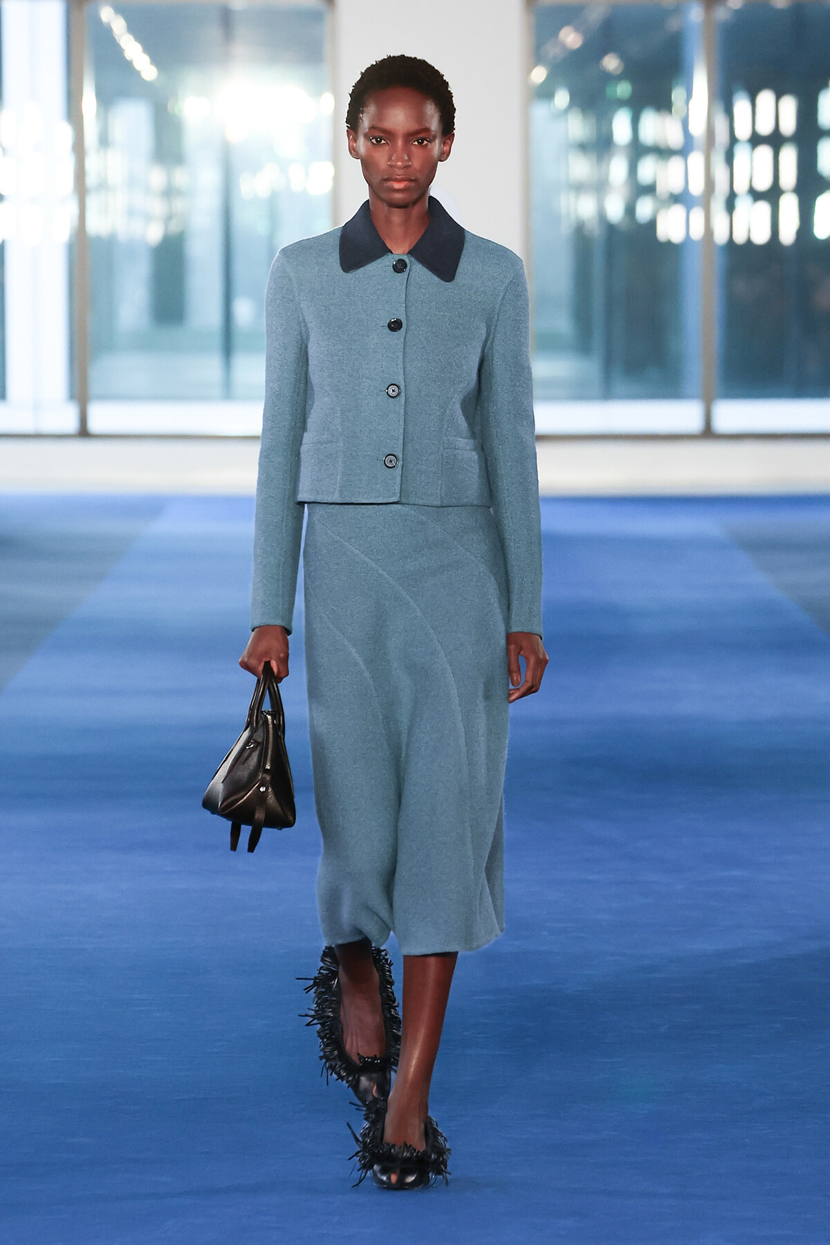 Model on a blue runway wearing a light blue cropped jacket with a dark collar and matching skirt, carrying a small black handbag.
